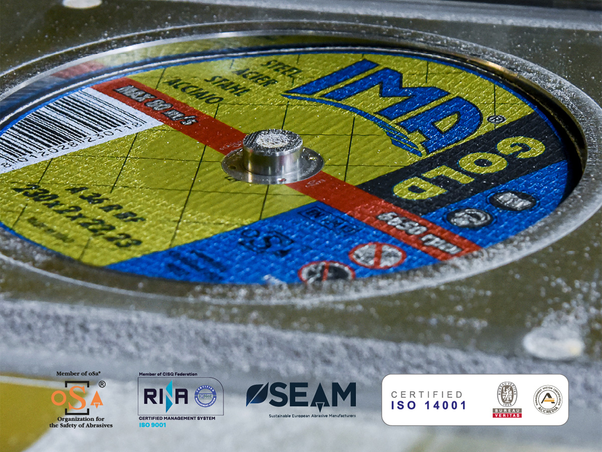 Foto di un disco abrasivo della line GOLD di IMA in fase di produzione. In basso tutti i loghi delle certificazioni che attestano la sicurezza dei prodotti (oSa), il sistema di gestione qualità dell'azienda (ISO 9001), l’iscrizione al programma di sostenibilità (SEAM, promosso dalla FEPA), l'integrazione della sostenibilità all'interno dei processi aziendali (ISO 14001), i requisiti di sicurezza per la progettazione e la produzione degli abrasivi (EN 12413).