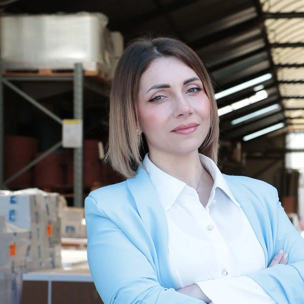 Foto ritratto di Francesca Aliberti, reparto commerciale della IMA Abrasivi. Bella ragazza con occhi azzurri e pelle chiara, gote rosate e capelli chiari, tra il castano chiaro e il biondo. Indossa una camicia bianca e una giacca di colore azzurro cielo. Ha le braccia conserte e accenna un sorriso. Sullo sfondo, il magazzino dell'azienda.